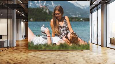 Two young adult woman having picnic in nature by the lake. Reading books, eating, having fun, enjoying holidays from school in summer. Wall mural