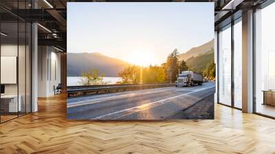 Big rig classic powerful semi truck transporting cargo in covered long bulk semi trailer running on the road along the river with mountain in sunset Wall mural