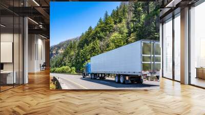 Classic blue American bonnet big rig semi truck transporting cargo in refrigerator semi trailer driving on the divided highway road with mountain and wild forest Wall mural