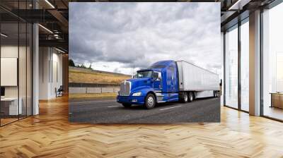 Classic blue big rig semi truck with dry van semi trailer going on wide highway with stormy sky Wall mural