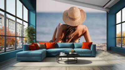 Young lady in a bathing suit and hat sits on the sand on the beach, looking towards the sea. Beautiful girl is facing the ocean with her back to the lens Wall mural