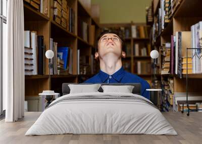 Guy in blue shirt standing in the library in a pose like praying to God, geasute of despair. Young student stressed due to excessive studies. Wall mural