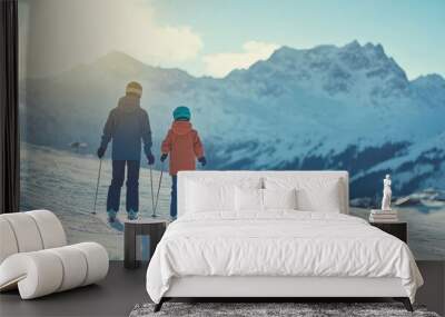 A snowy landscape features two skiers, an adult and a child, standing on the slope. They are enjoying a sunny day, surrounded by majestic mountains Wall mural