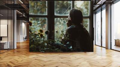 A woman sits quietly by the window, gazing at sunlit flowers and greenery in a peaceful garden during the late afternoon Wall mural