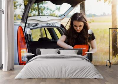 Young attractive girl stands near car with open back door and emergency lights is on, tries to find something in first aid kit. Concept of car crash and giving first aid Wall mural