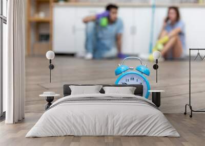 Close up photo of alarm clock and tired couple after exhausting cleaning day sitting on the floor and resting on background. Time to household concept Wall mural