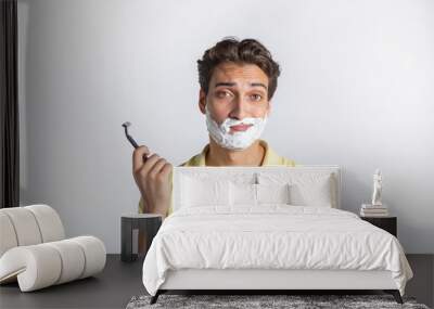 Close-up portrait photo of handsome man with a shaving foam on his face and holding razor blade isolated on white background Wall mural