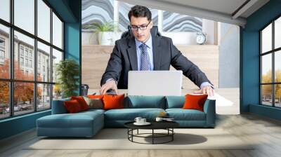 Handsome serious businessman in black suit sitting at the desk and working on laptop in the modern office	 Wall mural