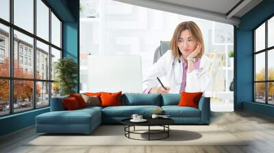 Portrait photo of young and beautiful female doctor in white medical gown sitting and working on laptop in the office of the modern clinic. Online consultation and distant cure concept Wall mural