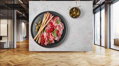 Slices of prosciutto coppa di parma ham with basil, grissini sticks, capers and redcurrant on dark plate on textured background. Top view with copy space Wall mural