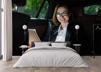 Successful attractive businesswoman sitting in the passenger seat in her luxury car working on laptop and driving to a business meeting Wall mural