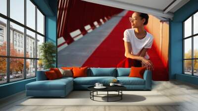 Bright summer lifestyle portrait of young pretty woman in eyewear, red skirt and white T-shirt, sitting on a bright red floor on the bridge. Wall mural