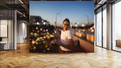 Bright summer lifestyle portrait of young pretty woman in eyewear, red skirt and white T-shirt, standing on the bright red bridge at evening with city background and blurred bokeh. Wall mural