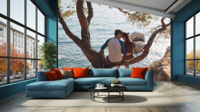 romantic young couple in love together. Happy young couple in love sitting and kissing on a tree branch on sea in background Wall mural