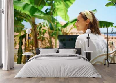 Young beautiful mixed race woman sitting on terrace at morning in hotel, using laptop computer while enjoying vacation Wall mural