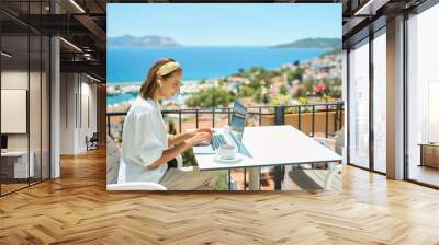 Young woman using laptop computer at cafe balcony of resort hotel with sea view, working typing emails browsing online enjoying drinking coffee Wall mural