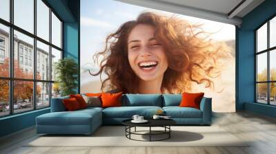 Beautiful young woman in gray shirt with curly red hair blowing in the wind, smiling brightly and laughing, enjoying on the beach as her hair is blowing in the wind. Wall mural