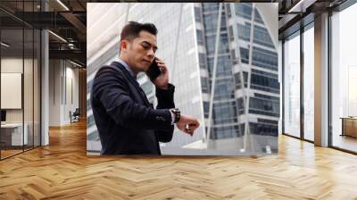 young smart asian business man wearing modern black suit looking at his watch and making phone call with mobile smart phone in city background, network connection, technology communication concept Wall mural