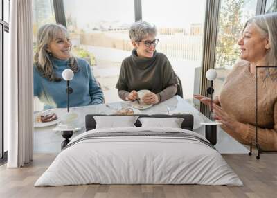 Three elderly women are sitting at a table in a coffee shop, talking and enjoying their tea. Wall mural