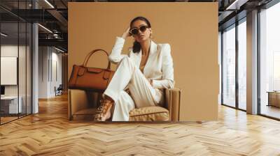 A chic woman in a white suit and round sunglasses sits confidently on a beige armchair, accompanied by a stylish brown handbag against a matching background. Wall mural