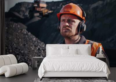 A construction worker with an orange safety vest and helmet listens intently through headphones amid a rugged mining site. Wall mural