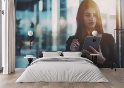 A focused woman in smart casual attire reads on a tablet, bathed in natural light within a modern office setting. Wall mural