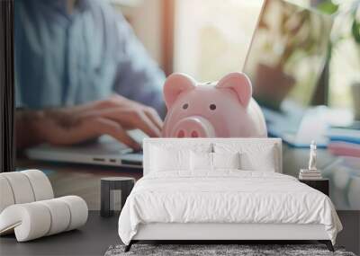 A person types on a laptop at a desk with a pink piggy bank in the foreground, symbolizing savings and financial planning. Wall mural