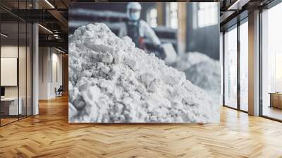 A pile of white powder sits in a factory, with a worker in protective gear standing in the background, highlighting industrial processes and safety. Wall mural