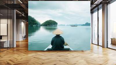 A serene scene of a person wearing a conical hat, sitting calmly on a boat, gazing at the tranquil waters and distant rocky islands under a cloudy sky. Wall mural