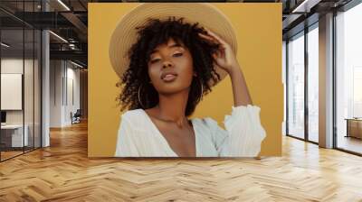 A stylish woman in a wide-brimmed hat and white blouse poses confidently against a vibrant yellow background. Wall mural