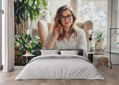 A woman in glasses and casual attire sits comfortably in a cozy armchair, surrounded by lush indoor plants and soft natural light in her home. Wall mural