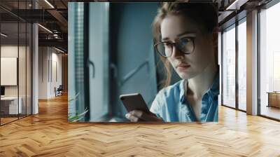 A young woman with glasses intently uses her smartphone by a window, surrounded by natural light and a potted plant, suggesting focus and modern lifestyle. Wall mural