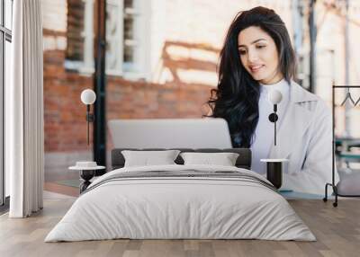 Adorable brunette woman in white clothes sitting outdoors at cafe using her laptop computer communicating with friends online using free internet connection. People, lifestyle, technology concept Wall mural