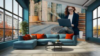 Beautiful businesswoman sitting on bench and working remotely on her project with laptop Wall mural