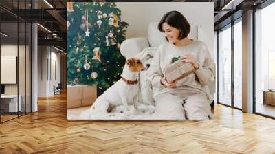 Cheerful brunette woman shows received presents to favourite dog, pose together on floor in cozy room, have festive mood, prepare for Christmas or New Year, enjoy winter time. Hoilday concept Wall mural