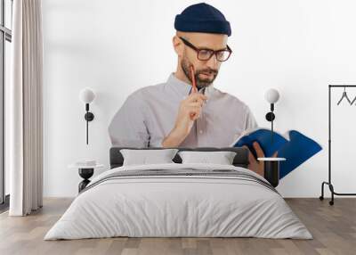 Concentrated unshaven adult man holds blue textbook and pencil, reads necessary information, has serious facial expression, wears formal clothes, isolated over pink background. Copywriter indoor Wall mural