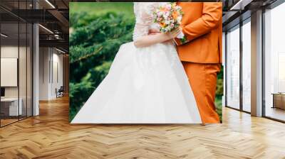 Cropped shot of young newlyweds pose against green nature background. Bride in white dress holds wedding bouquet stands next to groom in orange suit. People, wedding and relationships concept Wall mural