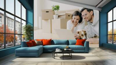 Glad young female and male discuss home repair projects, look attentively into laptop, their dog lies near, pose in living room on floor, dressed in casual clothes. People and new home concept Wall mural