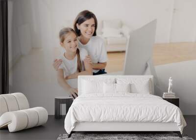 Happy brunette mother helps child do homework on computer, sit together at workplace, have cheerful expressions, focused into monitor. Little girl plays video games via modern gadget with mom Wall mural