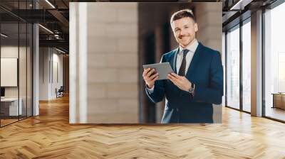 Happy male entrepreneur wears formal black suit, holds modern tablet computer, checks financial news in networks, connected to wireless internet, prepares financial report on business meeting Wall mural