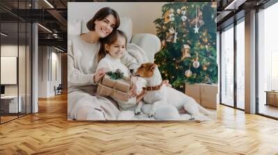 Happy winter holidays! Positive brunette woman embraces little girl pose with gifts on floor in room, jack russell terrier dog near, have fun near Christmas tree. Mom and daughter unpack boxes Wall mural