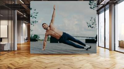 Healthy man stands in abs side plank and raises arm, does exercises outdoor, has muscular body and pleased expression. Handsome athlete trains in open air. Male couch balances on hand. Sport concept Wall mural