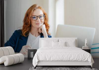 Horizontal shot of pleasant looking successful professional female lawyer learns clients case, works on modern laptop computer, dressed in formal apparel and transparent glasses, poses in office Wall mural