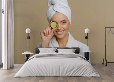 Horizontal shot of pleased refreshed woman covers eye with slice of fresh cucumber cares about skin uses organic products smiles pleasantly dressed in bath dressing gown after taking shower. Wall mural