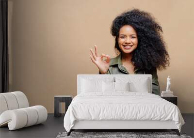 Indoor shot of pleasant looking curly woman has pleasant smile, makes okay gesture, excellent sign, gives approval, dressed in fashionable leather shirt, isolated over brown wall, blank space on left Wall mural