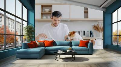 People, food, drink and pets concept. Horizontal shot of handsome young man eats tasty sweet pancakes, his pedigree dog looks with temptation, spend weekend at home, pose against kitchen interior. Wall mural