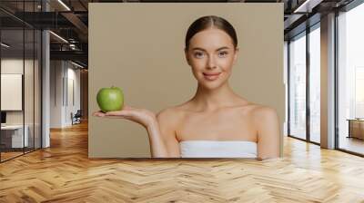 Photo of healthy lovely woman with natural makeup stand wrapped in bath towel holds green apple recommends product for dieting isolated over brown background makes natural homemade facial masks Wall mural