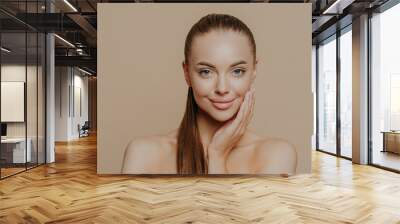 Photo of lovely young woman has smooth healthy skin after beauty procedures and receiving facial massage, stands bare shoulders, has long dark straight hair, isolated over beige wall cares of her body Wall mural