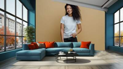 Photo of slim cheerful African American woman with curly hair, smiles happily, being in good mood, wears white t shirt and jeans, keeps hand in pocket, has slim figure, isolated on beige background Wall mural