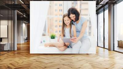 Portrait of pretty female embraces her little daughter, watch cartoon on digital tablet, connected to wireless internet at home, sit on window sill with white curtains at home. Children and parents Wall mural